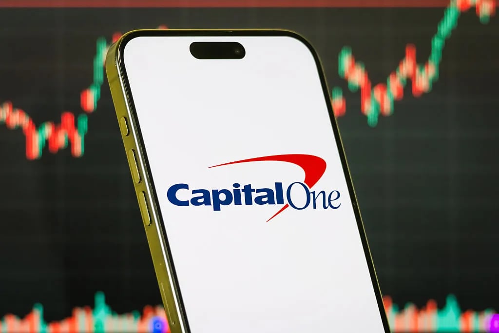 In this photo illustration, a smartphone displays the logo of Capital One Financial Corporation (NYSE: COF), a major American bank holding company specializing in credit cards, auto loans, banking, and savings accounts, in front of a screen showing the company's latest stock market chart on July 19, 2025 in Chongqing, China.