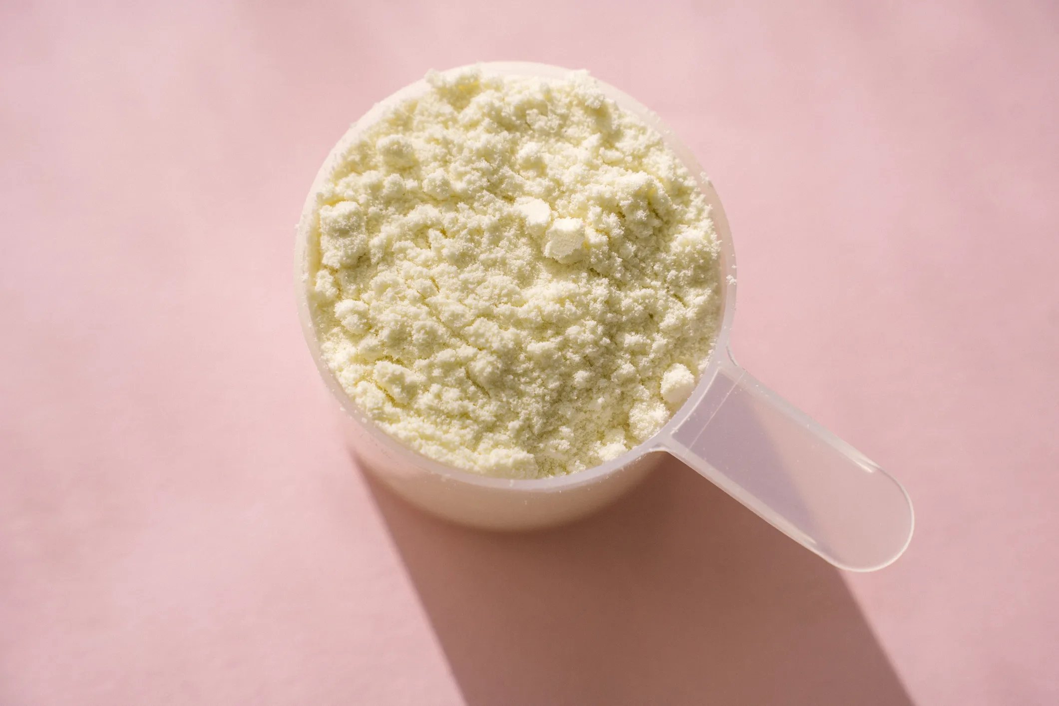 Measuring scoop of whey protein powder on pink table in bright sunlight