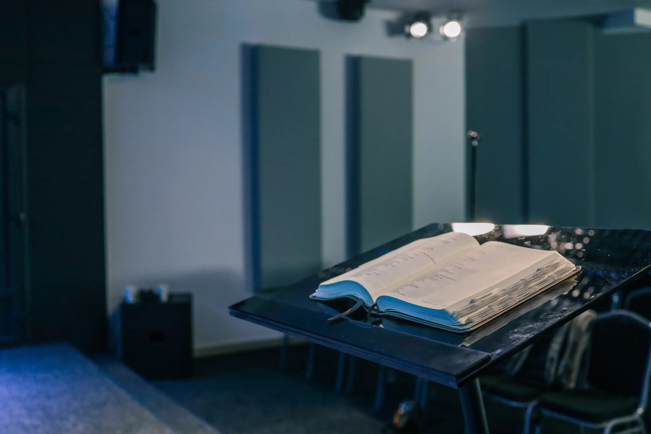 Open Bible resting on a black podium inside a modern church hall, with chairs and musical instruments visible in the background.