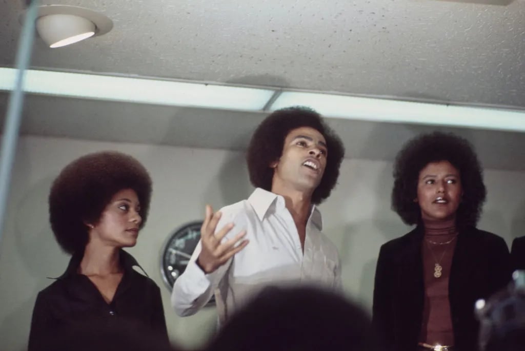 Black Panther Party co-founder Huey Newton (centre, 1942 - 1989) speaking to a crowd of circa 500 Black Panthers on arrival at San Francisco International Airport, California, July 5th 1977. He is with his wife Gwen (left) and Black Panther chairwoman Elaine Brown.