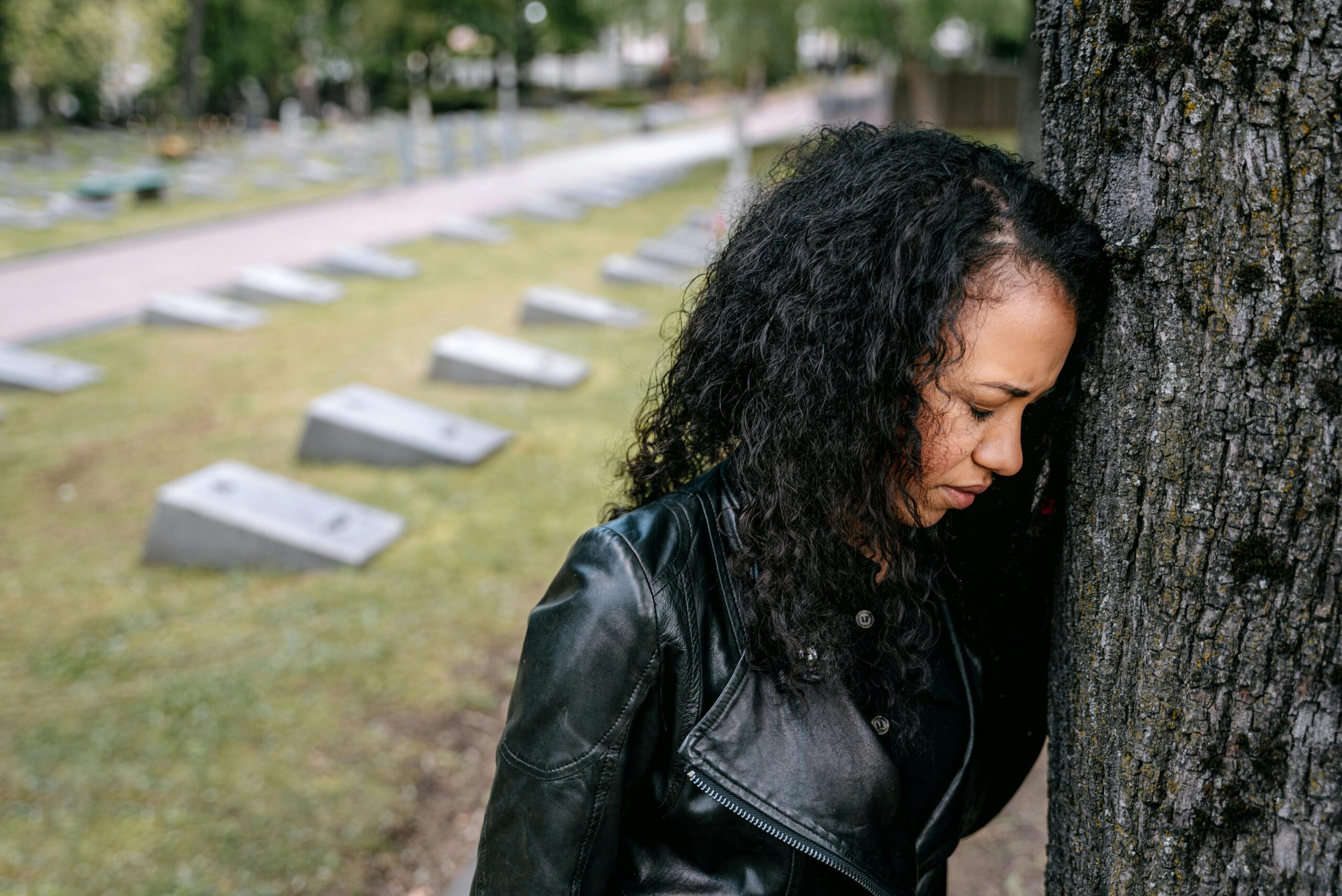 A Black woman in a black leather jacket reflects solemnly against a tree in a cemetery.