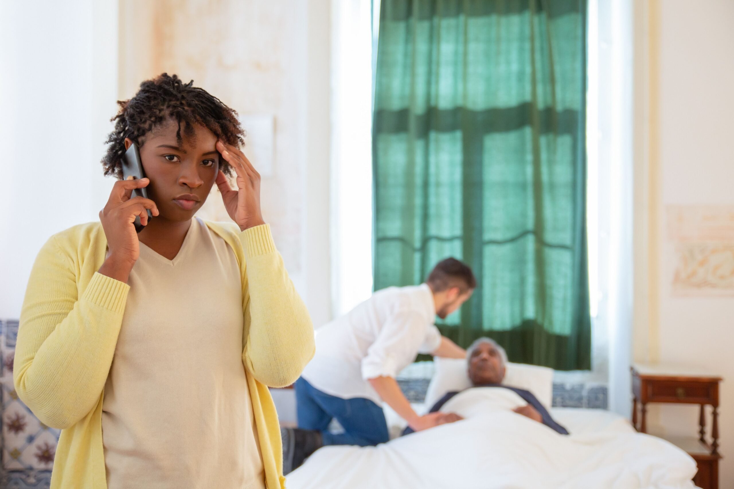 A Black woman on the phone looks worried as a caregiver assists an elderly person in bed.