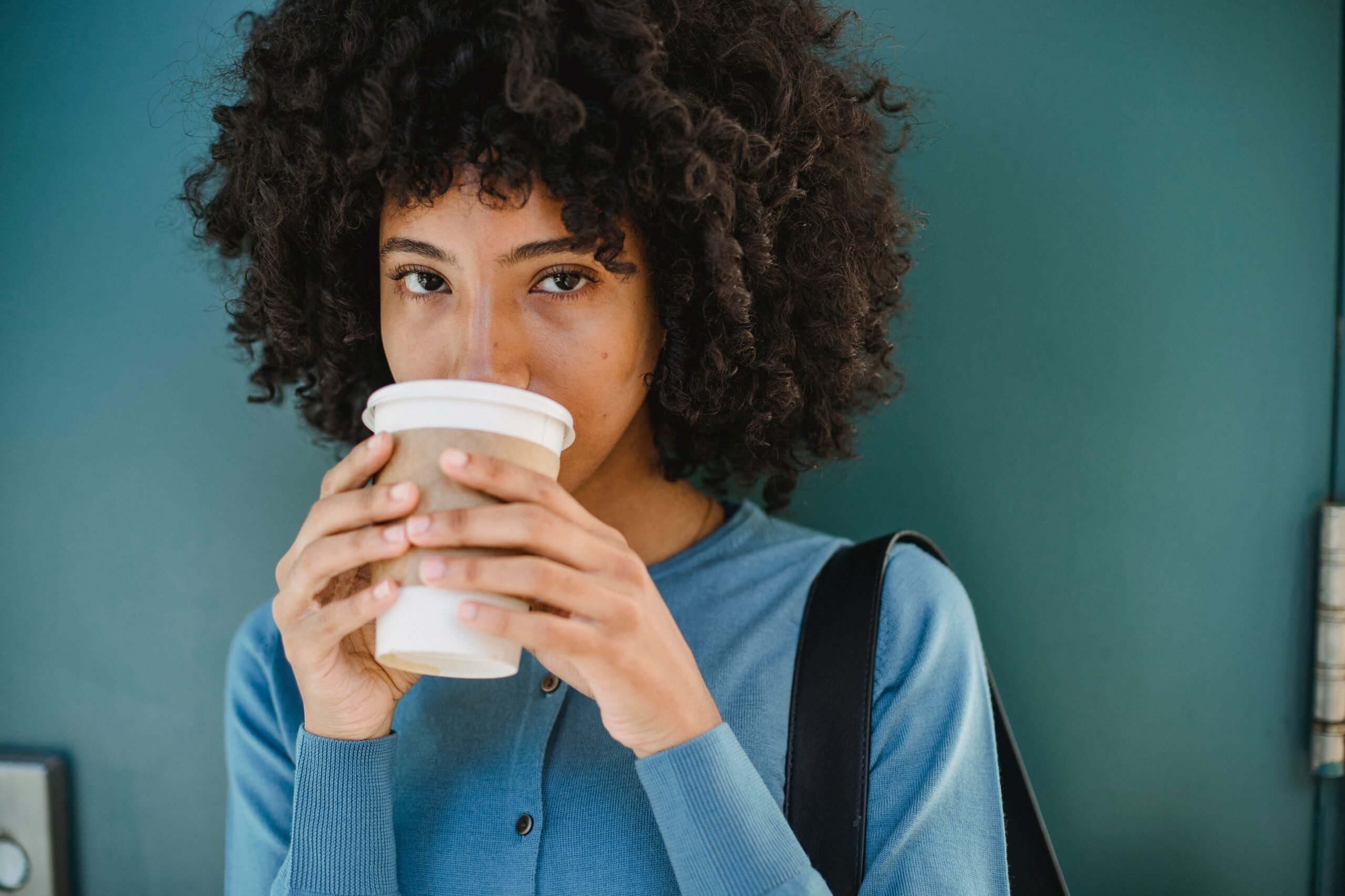 Portrait of a Black woman savoring a hot drink with a teal backdrop, casual and relaxed.