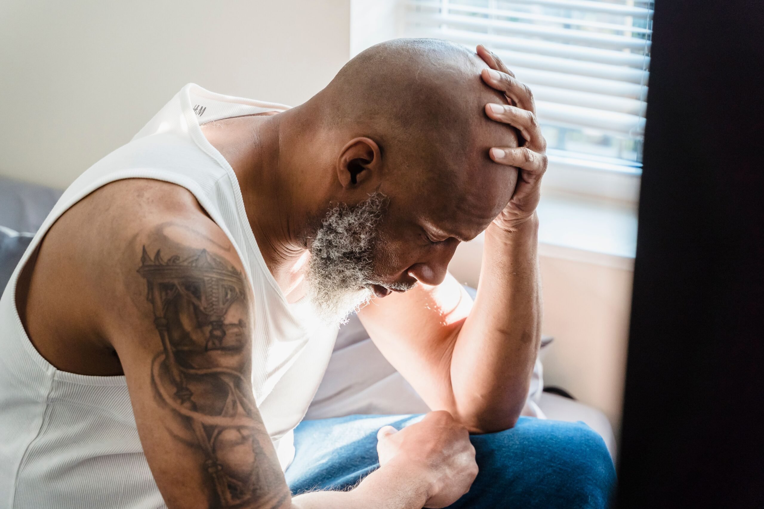 A Black senior man sits thoughtfully at home, conveying emotions of contemplation and stress.