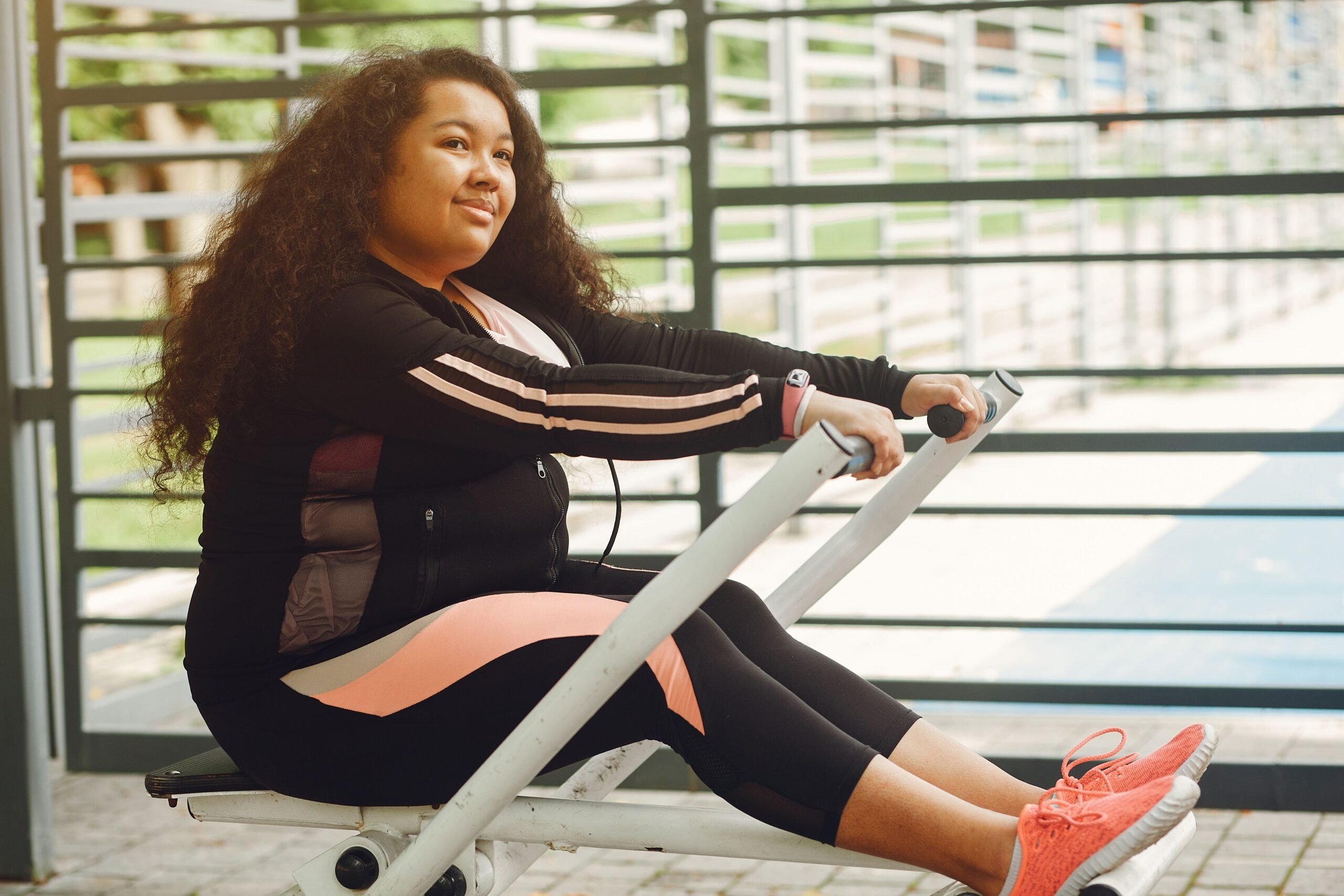A plus size woman smiling and working out on a rowing machine outdoors.