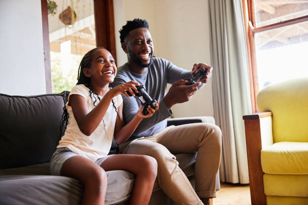 70s trivia Pictured: Little girl and her father laughing while playing video games together on a sofa in their living room at home