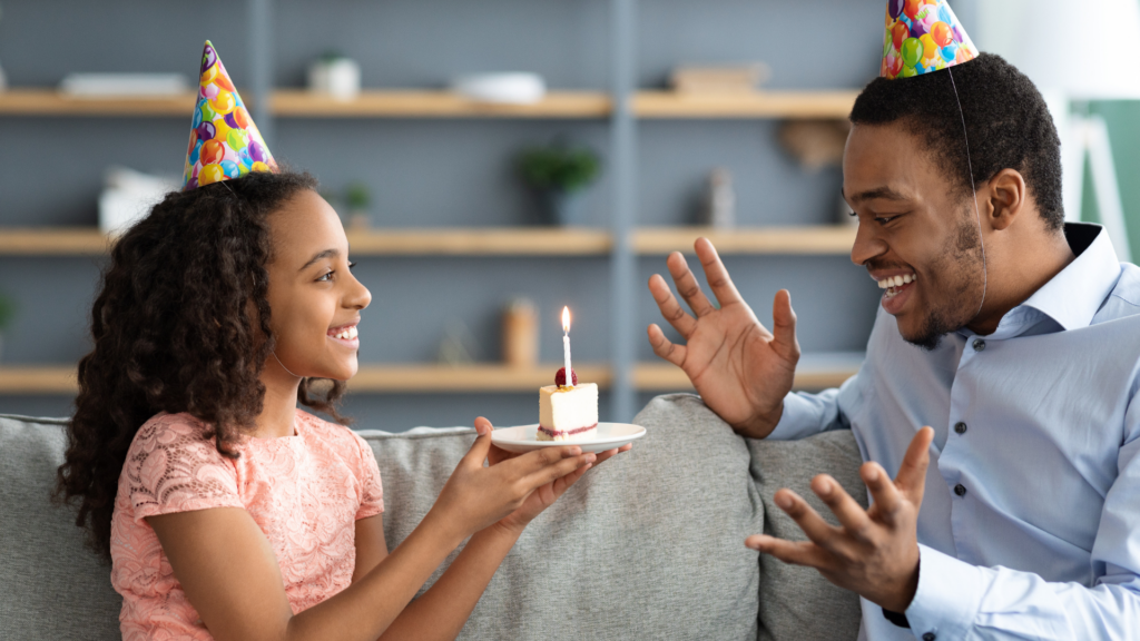 Beautiful black girl holding birthday cake  with lit candle for her excited dad wearing nice outfits and part hats african american father and daughter celebrating birthday together at home