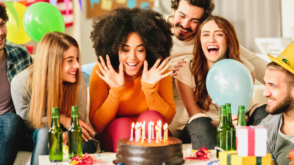 Group of friends celebrating a birthday with beer and chocolate cake on a table surrounded by colorful party balloons 