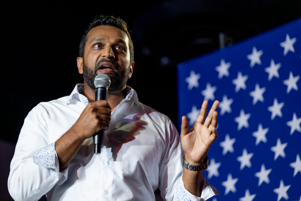 Trump Administration member Kash Patel holding a mic with an American flag behind him