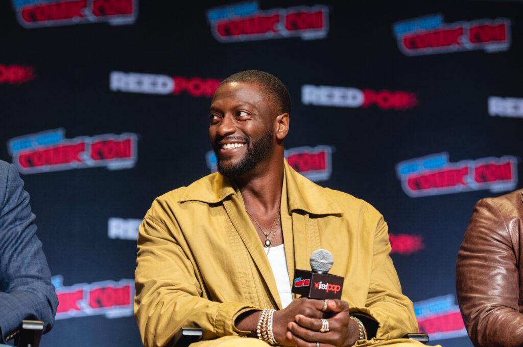 Aldis Hodge at a premiere event sitting on stage and holding a mic while wearing a tan jacket and white shirt